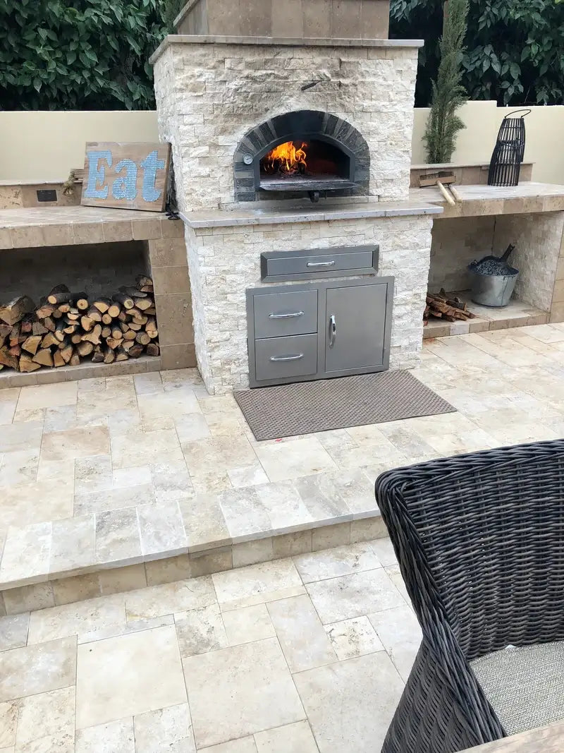 Pizzaioli Stone Arch built into a stacked travertine outdoor kitchen wall with the fire burning and firewood stored below