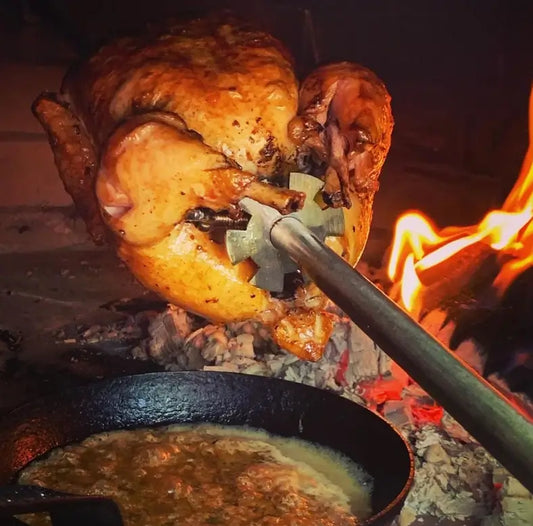 Whole chicken on the Authentic Pizza Ovens rotisserie spit turning beside a wood fire in a brick oven, drippings caught in a cast iron pan below