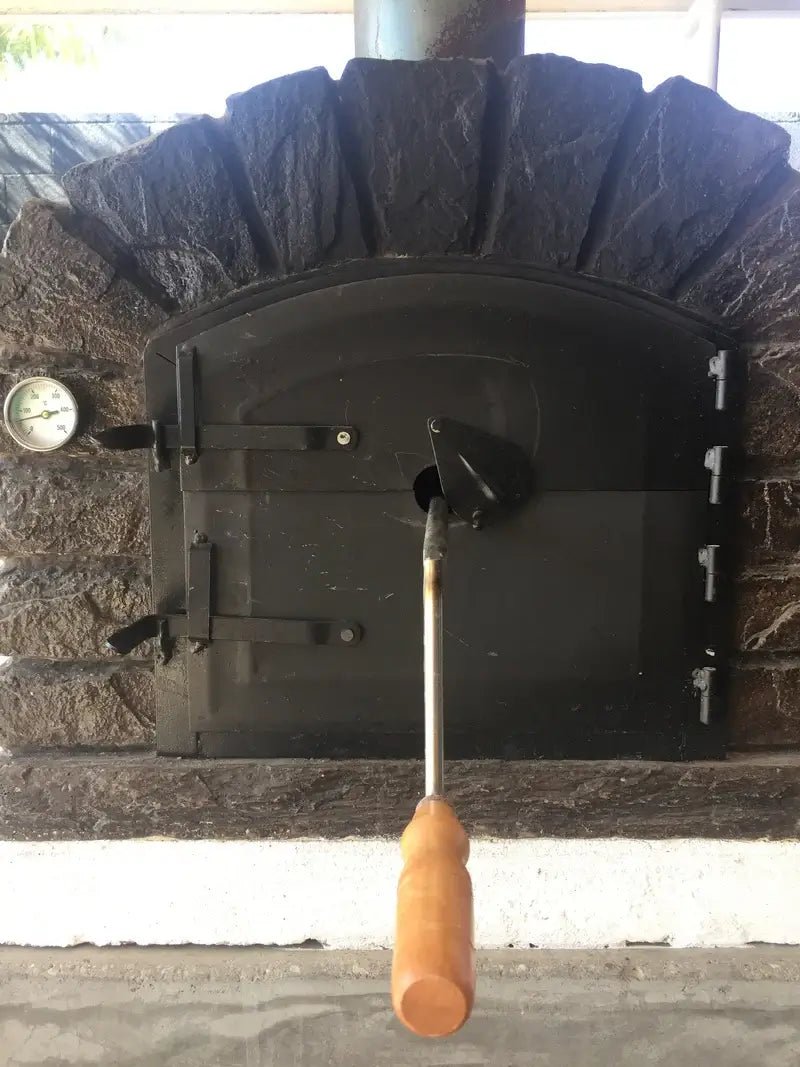 Wooden handle of the Authentic Pizza Ovens rotisserie spit passing through the spit port in the cast aluminum door of a stone arch oven