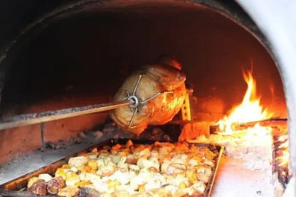 Roast turning on the Authentic Pizza Ovens rotisserie spit over a tray of potatoes and vegetables in a wood fired brick oven