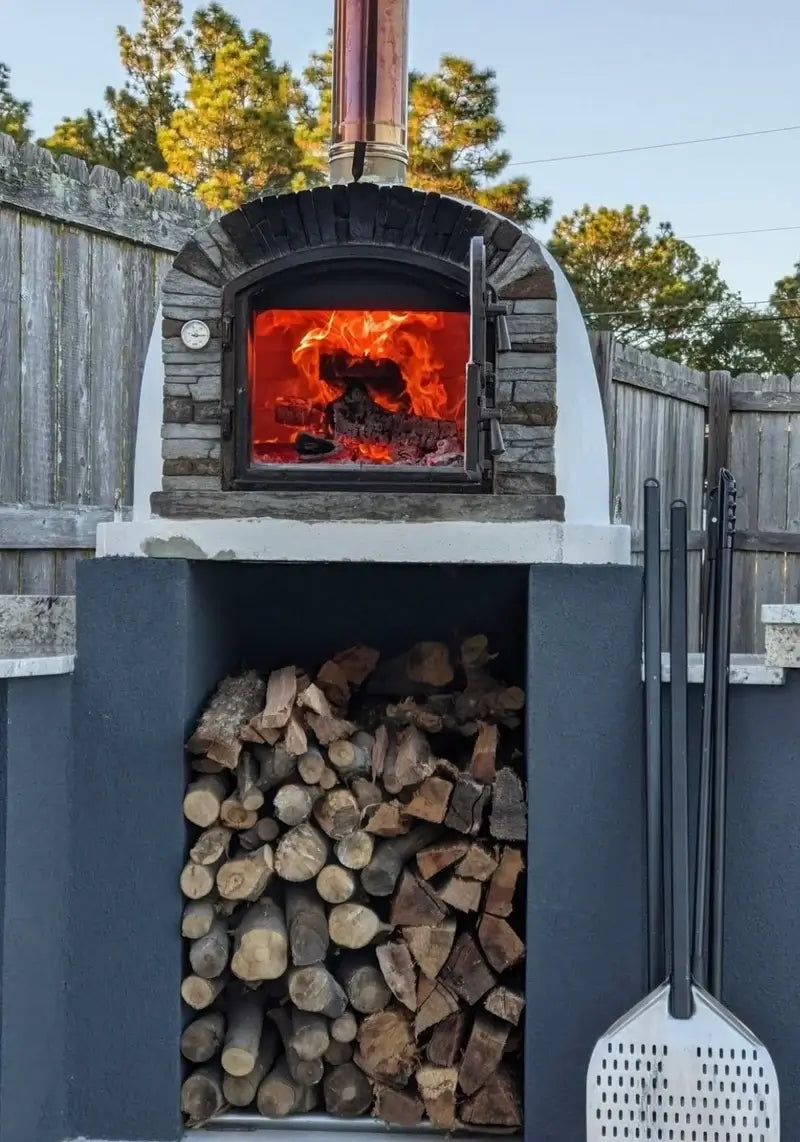Ventura Preto pizza oven with a full fire burning, installed on a dark stucco base with firewood storage and pizza peels alongside