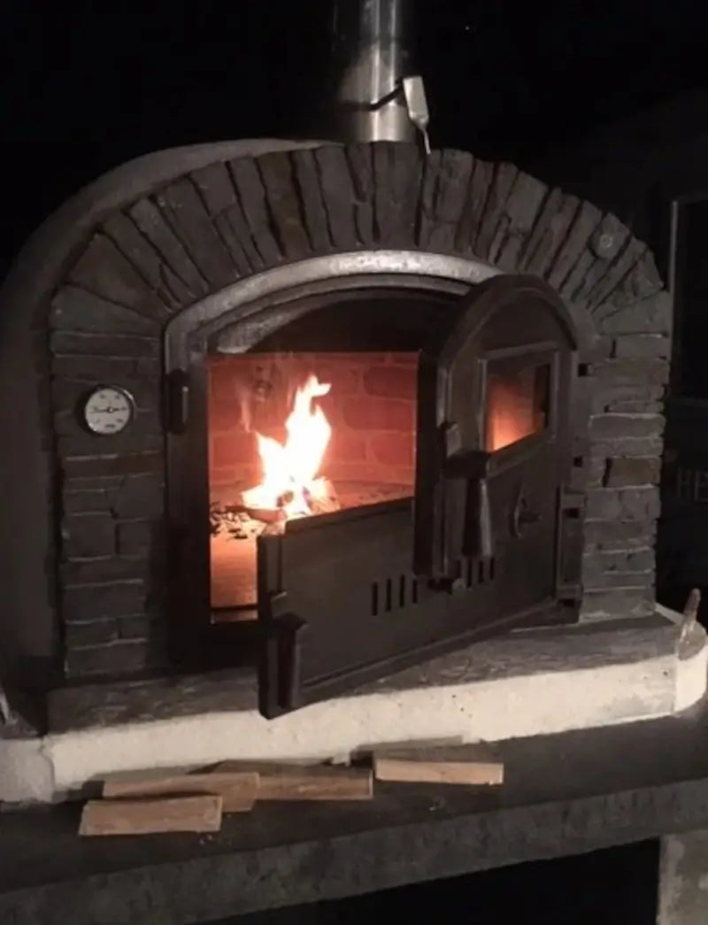 Ventura Preto pizza oven at night with one half of the Dutch door open and flames lighting the fire brick chamber