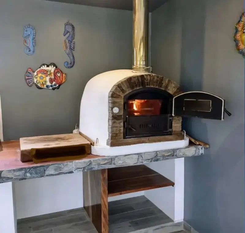 Ventura Sierra pizza oven with a fire burning and the door open, built into a stone topped counter in a covered outdoor kitchen with a wood peel alongside