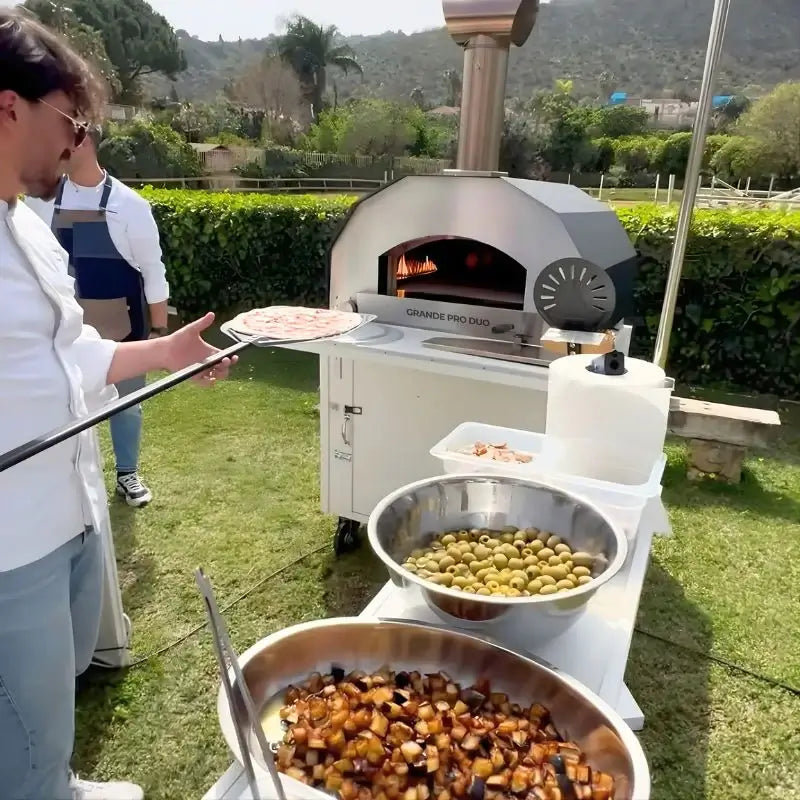 ItaliaForno Grande Pro Duo on its cart outdoors with a cook holding a topped pizza on the peel